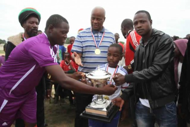 CULT HERO . . . Dynamos midfielder Ronald “Rooney” Chitiyo (right) helps young Mwalimu Kumbula to hand the winners’ trophy to Eddington Kuyaupi (left), the captain of Liverpool, while the event’s sponsor Musekiwa Kumbula (centre) looks on at Kubatana Primary School on Sunday
